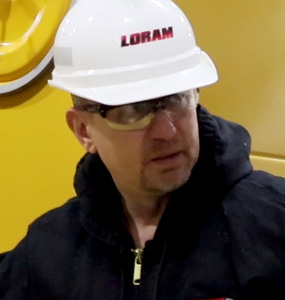 A man wearing a hoodie, safety glasses, and a Loram hard hat stands in front of a yellow background