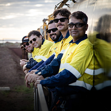 A group of men wearing yellow and blue jackets and sunglasses lean against a railing and smile.
