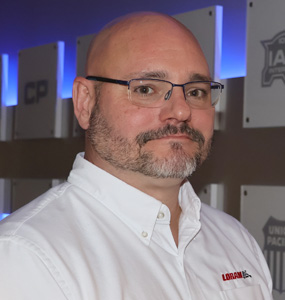 A man with glasses and a white LORAM button down shirt stands in front of a wall with white placards