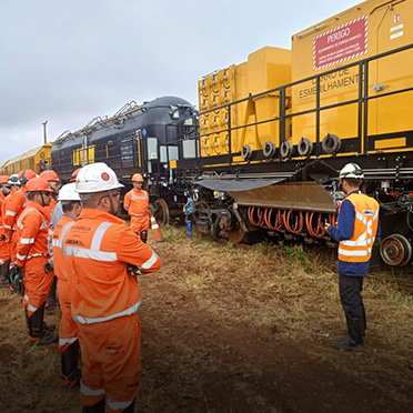 A line of men in orange reflective suits and hard hats stands in front of a train outside, listening to a man in an orange vest and white hard hat.