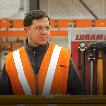 A man in an orange reflective vest stands outside in front of a Loram building